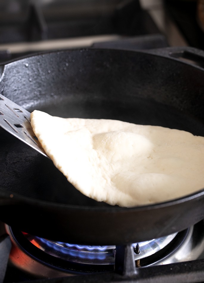 Een close-up van een metalen spatel die een stuk naanbrood in een koekenpan optilt