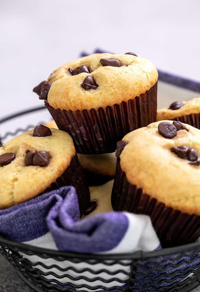Pile of chocolate chip muffins in brown liners in a wire basket lined with blue and white striped towel