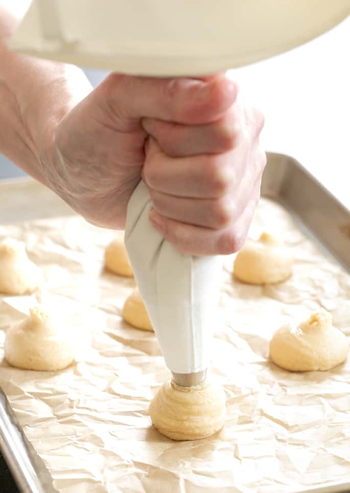 Gluten free nilla wafers raw batter being piped from a white piping bag onto a baking sheet with brown paper