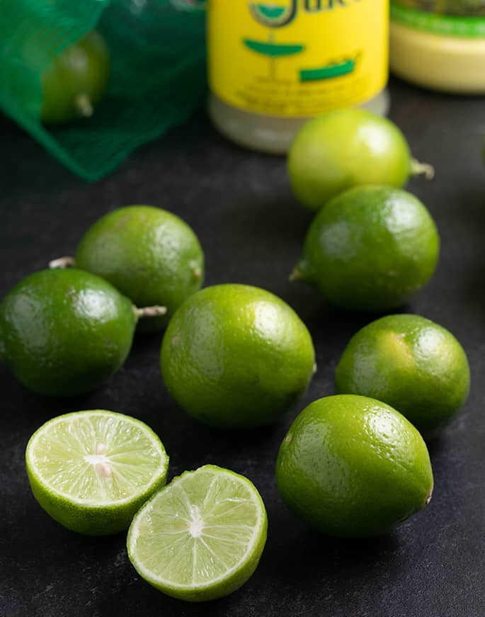 Key limes on black table with one sliced in half and bottles in background