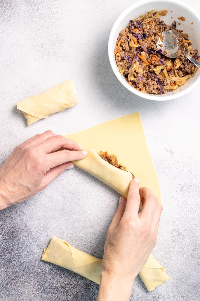 Overhead image of hands folding egg roll wrapper over filling with bowl of filling and more shaped egg rolls