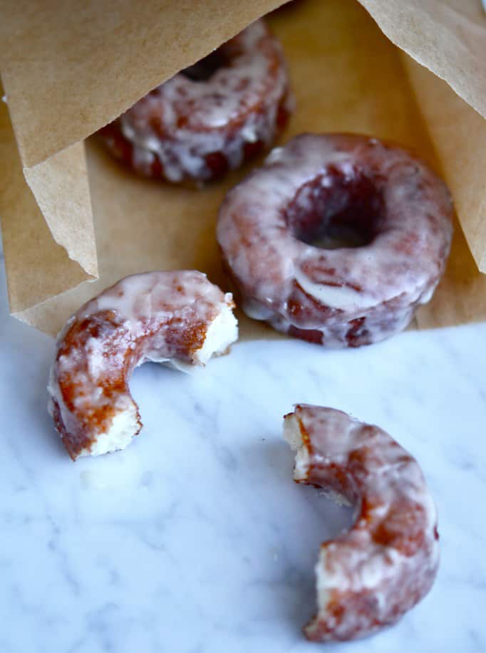 3 glazed donuts coming out of a paper bag onto a marble suface, one donut broken in half