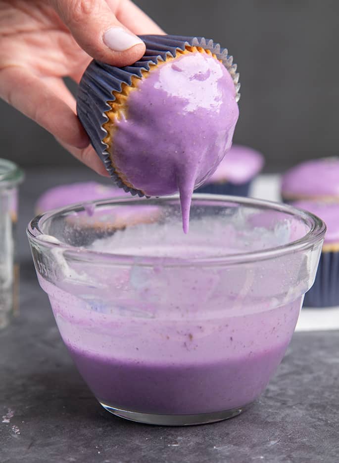 Fingers holding cupcake with dripping lavender glaze over glass bowl