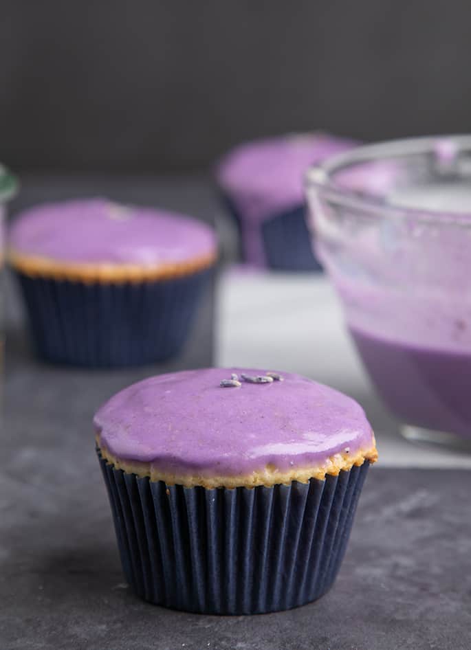 Yellow cupcake with purple glaze and lavender flower buds on gray surface