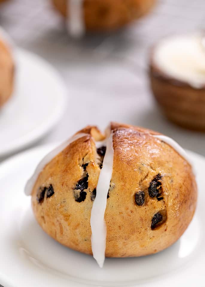 Hot cross bun with frosting cross on small white plate with butter in background