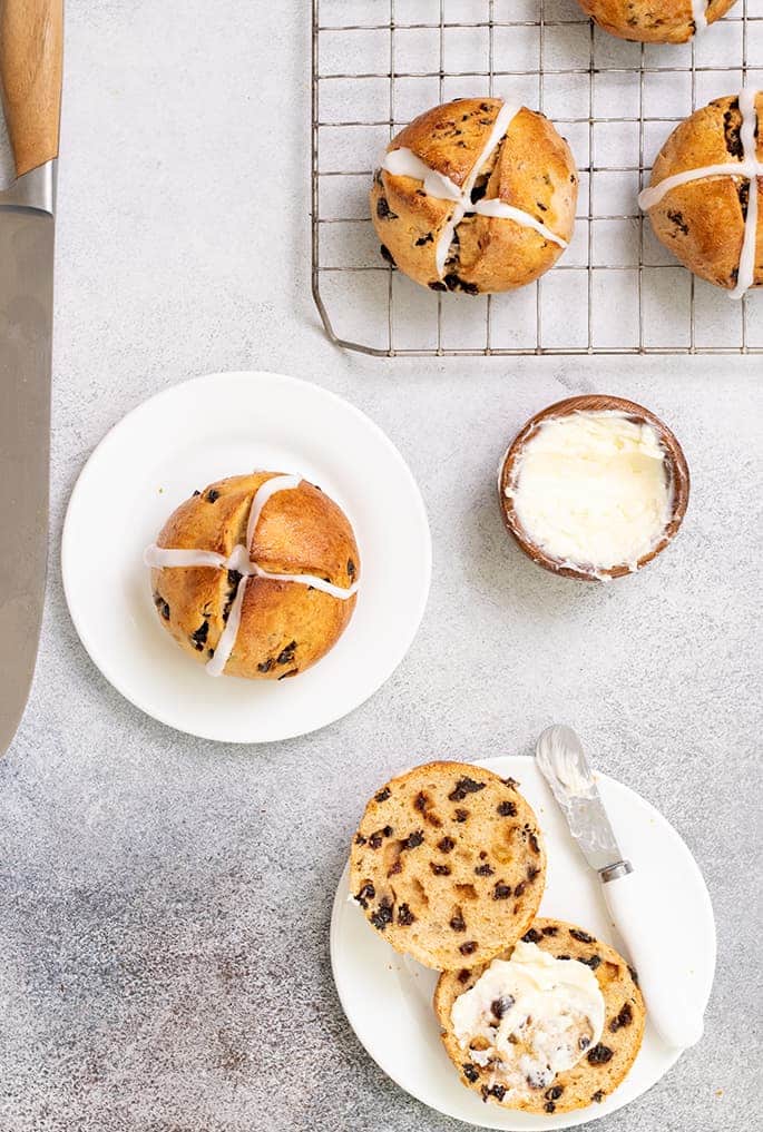 Overhead image of large knife, hot cross buns on wire rack, on small plate, and on small plate cut in half with butter