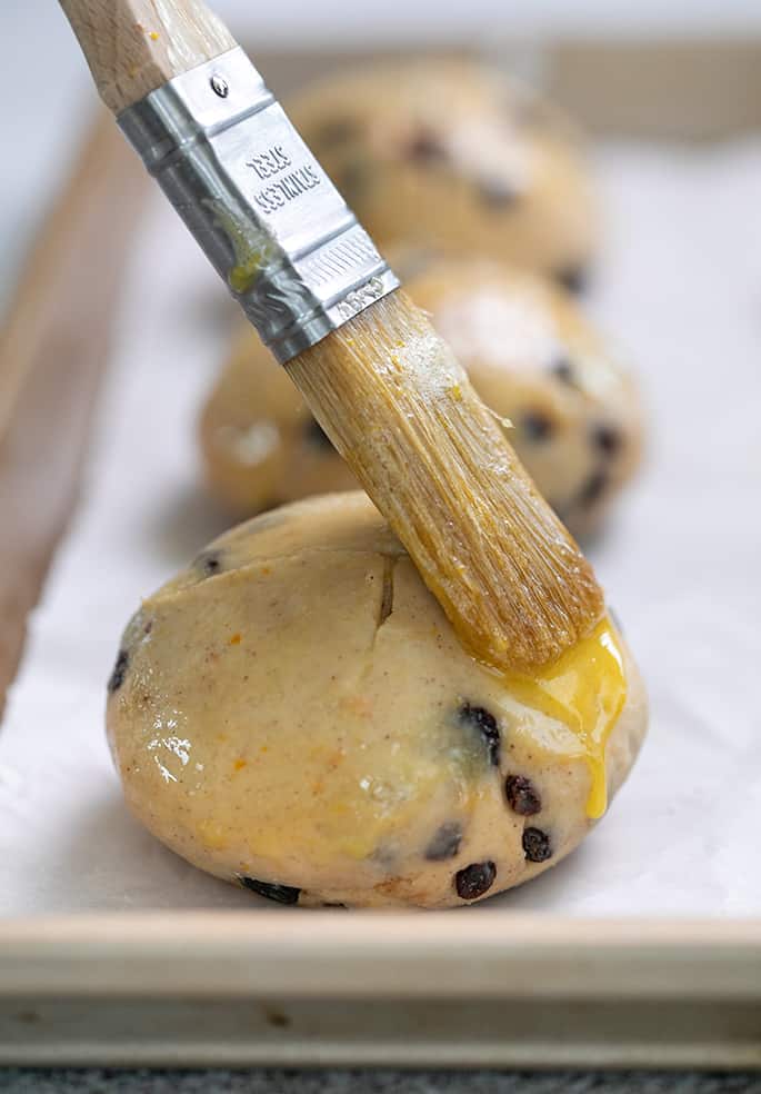 pastry brush placing glaze on risen hot cross bun on white paper on tray