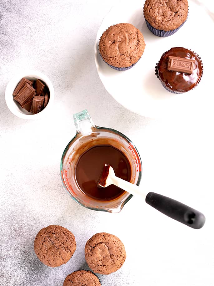 Array of chocolate glaze in glass measuring cup, some glazed cupcakes and some unglazed