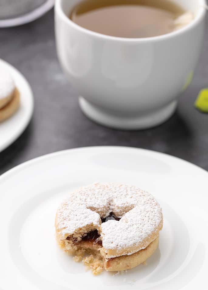 Broken linzer cookie on small white plate with cup of tea in background