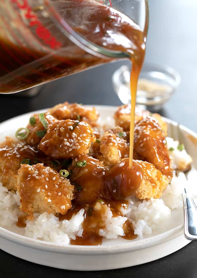 Gluten free chinese food recipe for sesame chicken on white rice on plate with more sauce being poured onto chicken