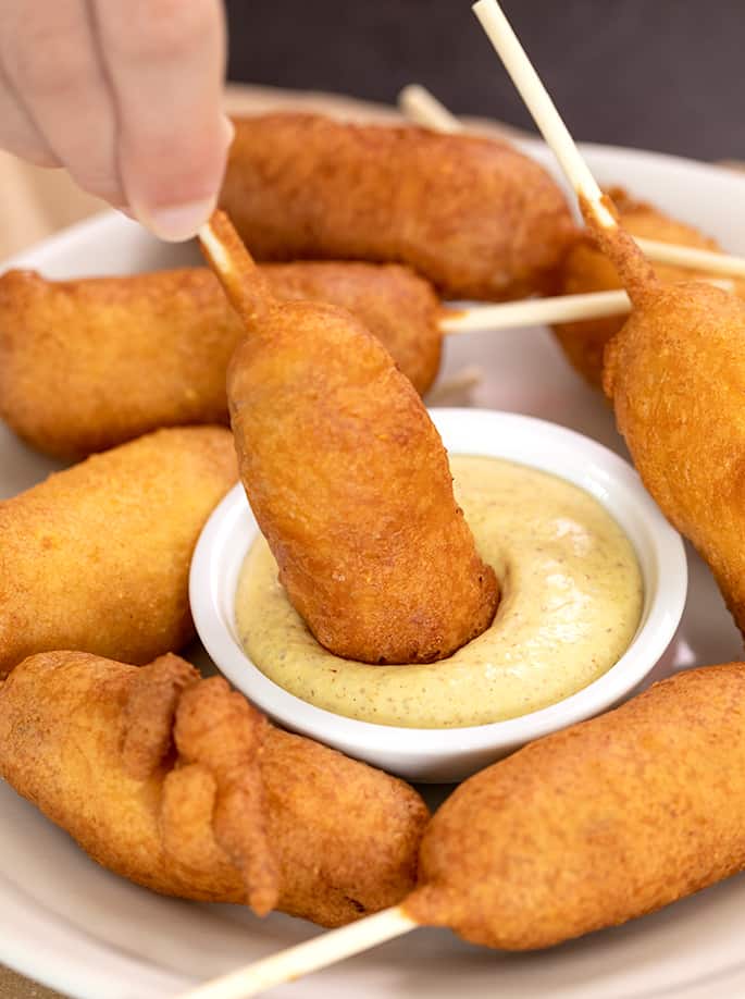 Hand holding stick of corn dog to dip it in small bowl of mustard