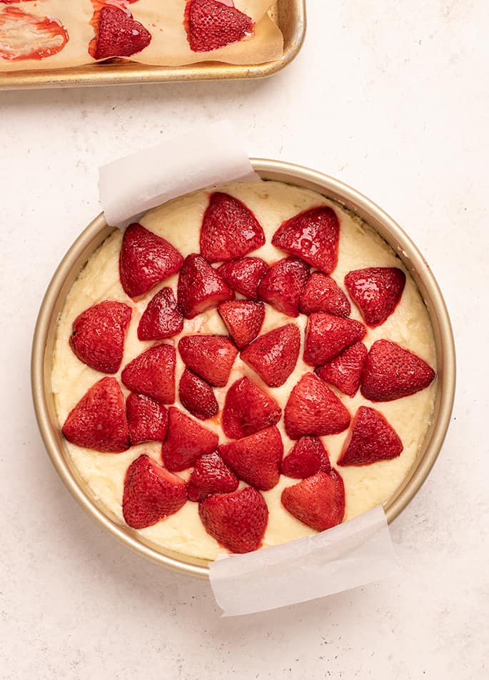 Overhead image of raw yellow cake with halved roasted strawberries on top in round baking pan with parchment paper