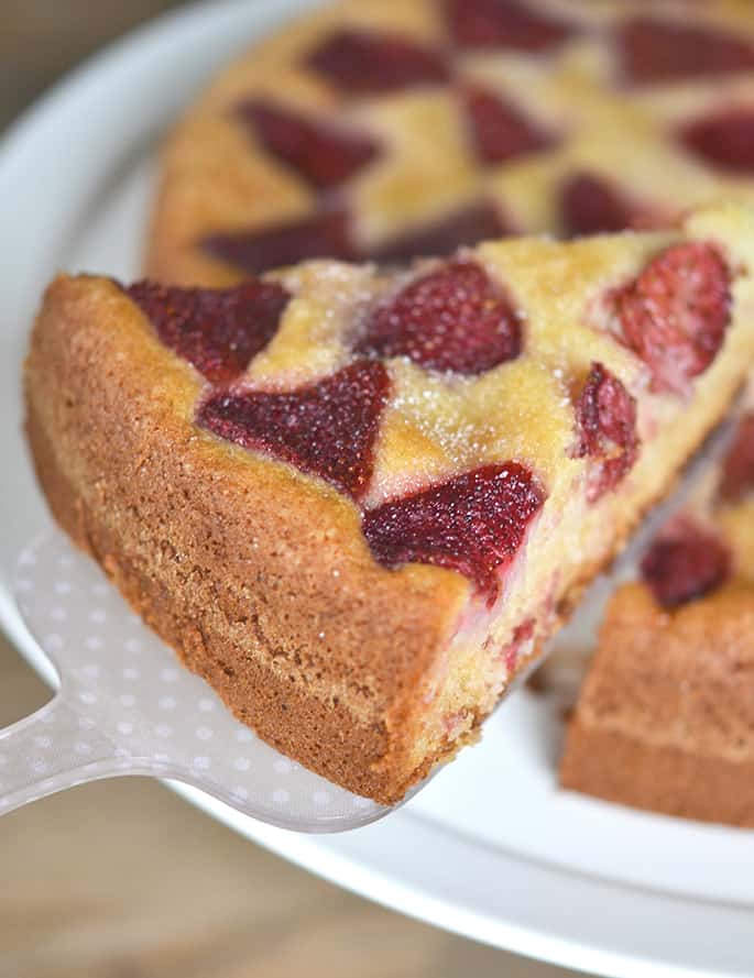 Slice of roasted strawberries on brown cake being lifted off white serving platter with rest of cake