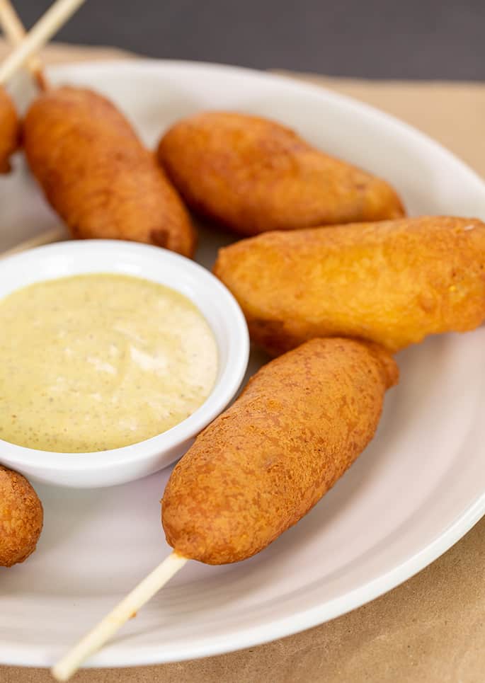 White plate with corn dogs arranged on top of brown paper and white stick in foreground