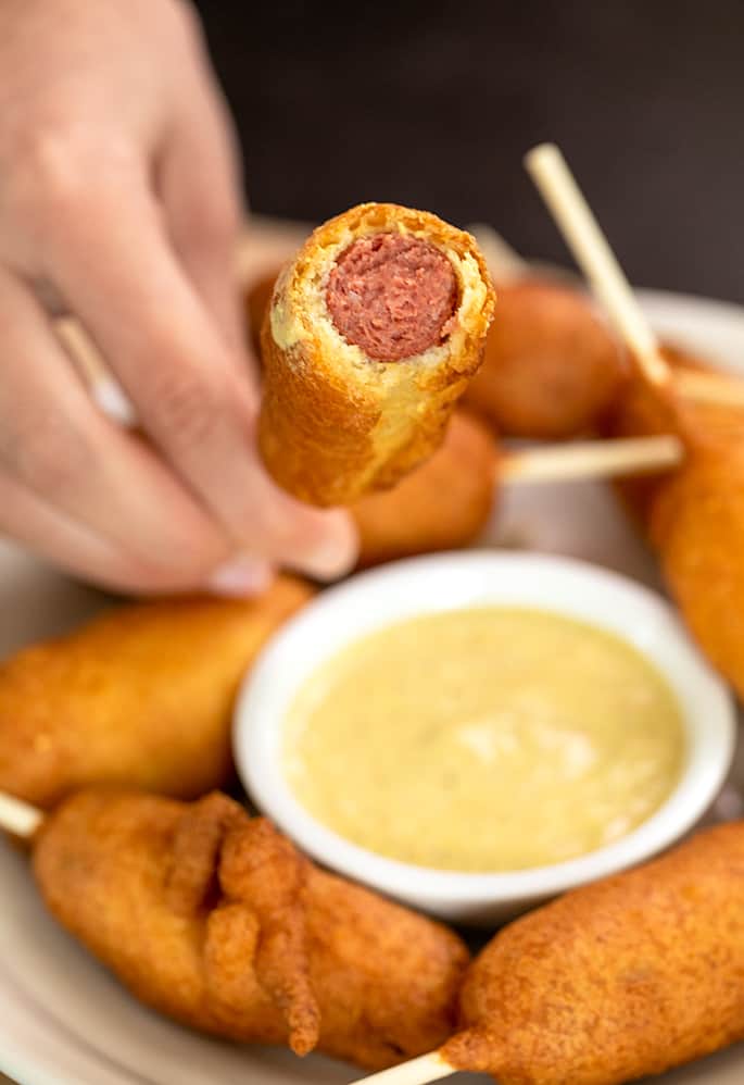 hand holding corn dog stick with end bitten off to reveal hot dog inside, over small bowl of mustard