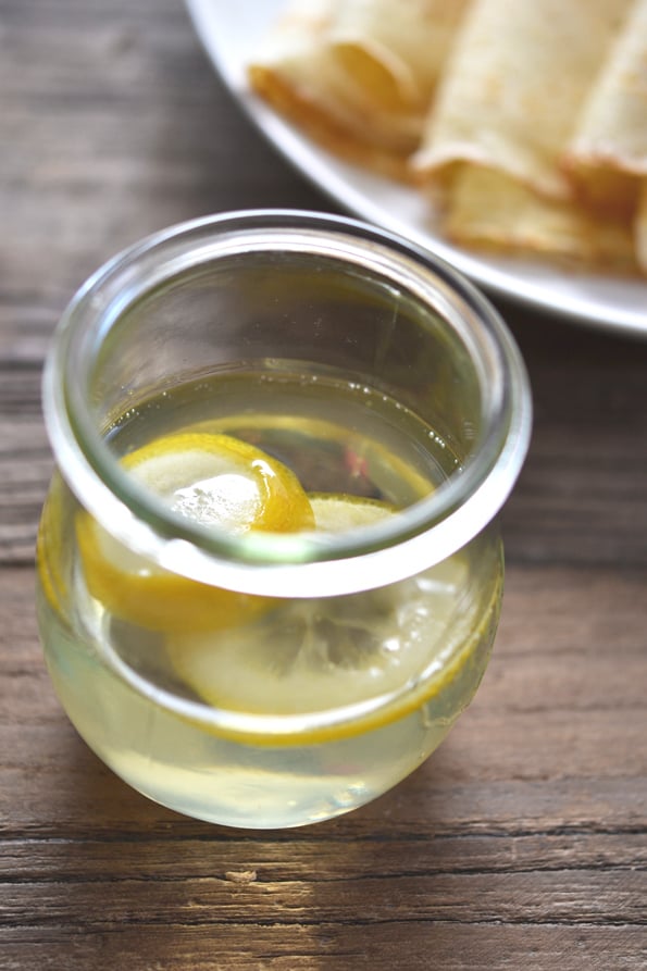 A cup of lemon water in a glass with a plate of crepes in background