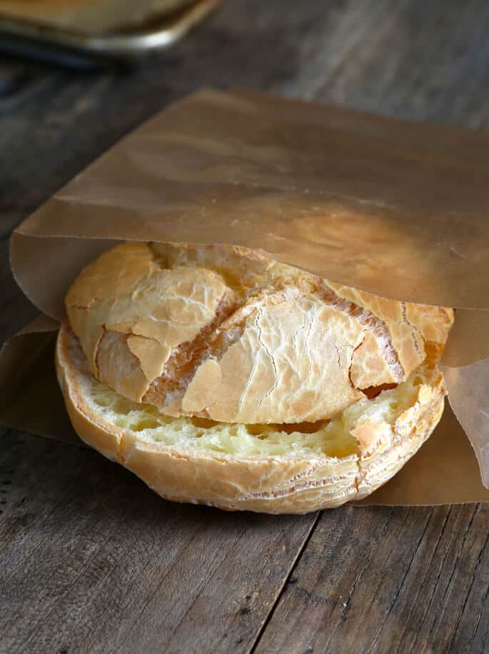 Sliced small round bread roll with very light brown crust and pale yellow inside with holes in a brown wax paper bag