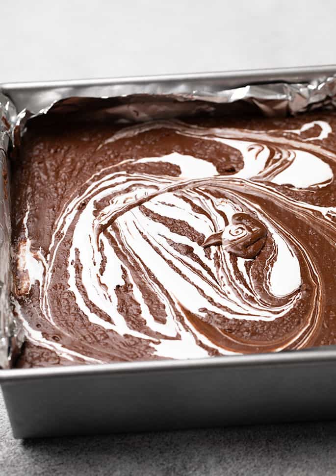 Brown fudge with white swirl on top in square metal pan lined with aluminum foil