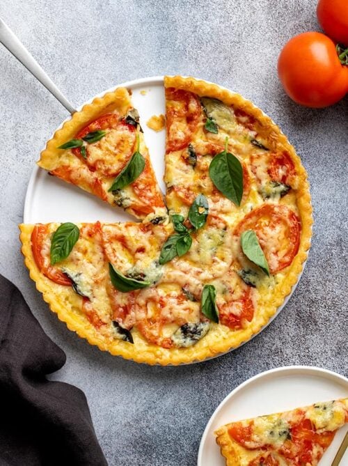 Overhead image of tomato tart on white serving plate with two traingular slices cut and one slice on a small white plate with whole tomatoes and a dark gray cloth