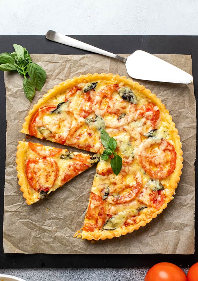 Sliced tomato tart on brown paper and a black board with pie server, basil leaves and tomatoes