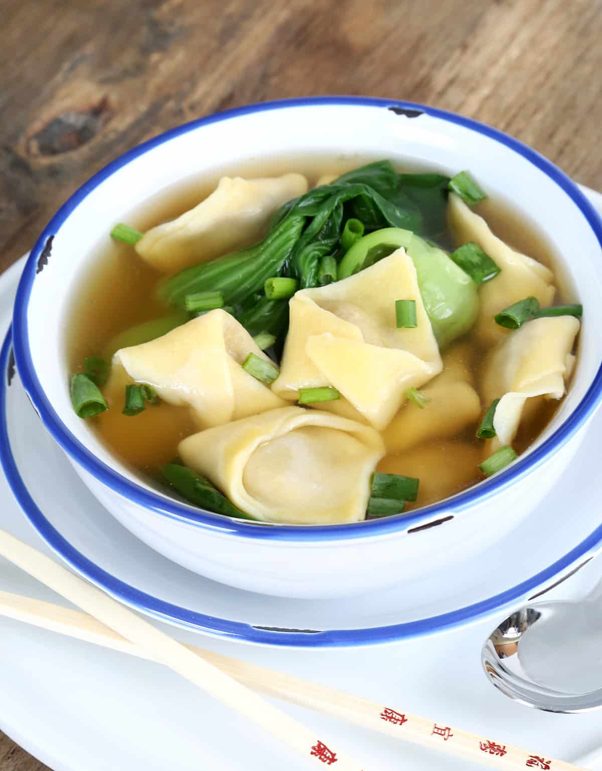 Light brown soup liquid with soup dumplings, chopped scallions, and baby bok choy in white bowl on white plate with chop sticks and a spoon