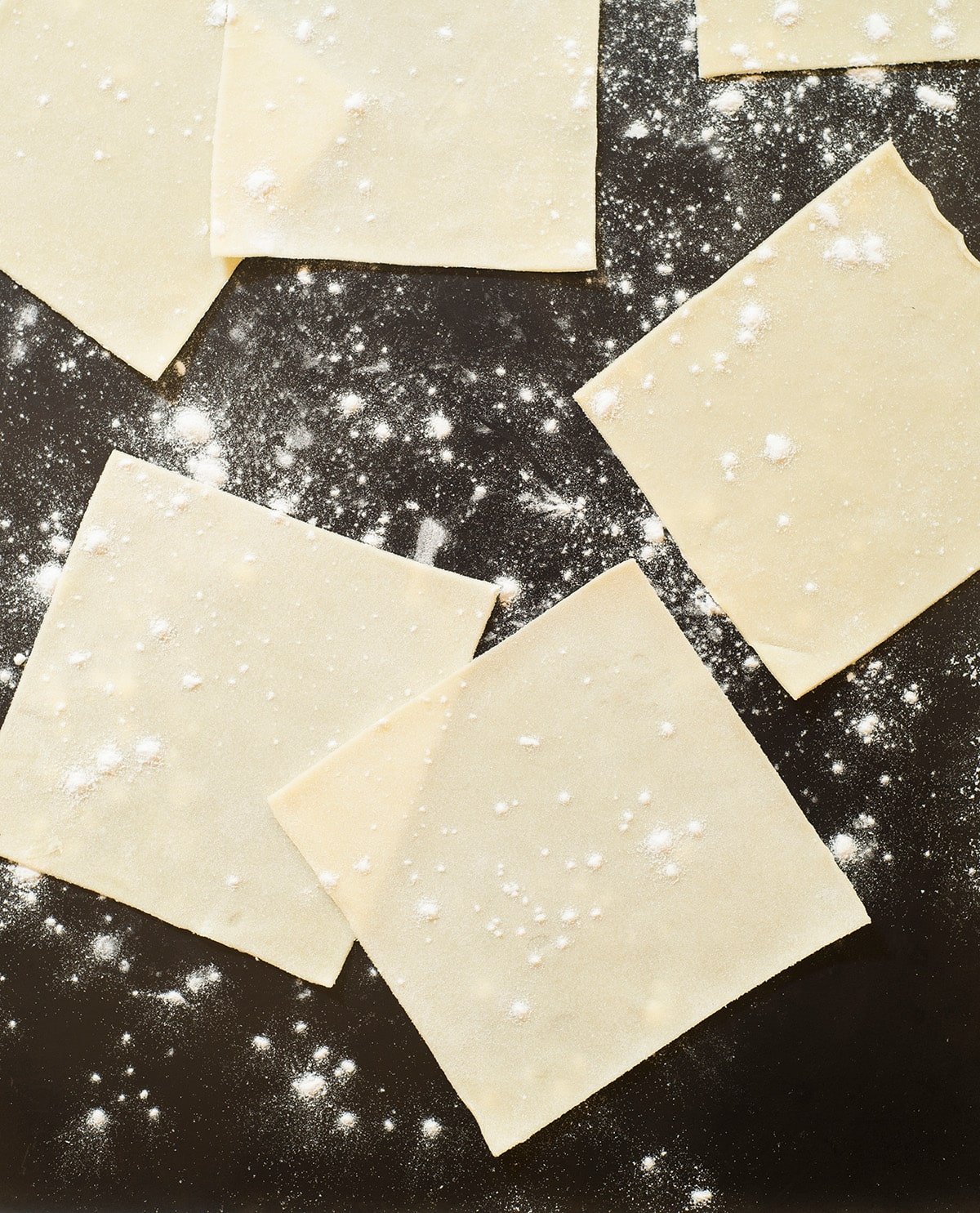thin yellow squares of dough with white flour on black surface