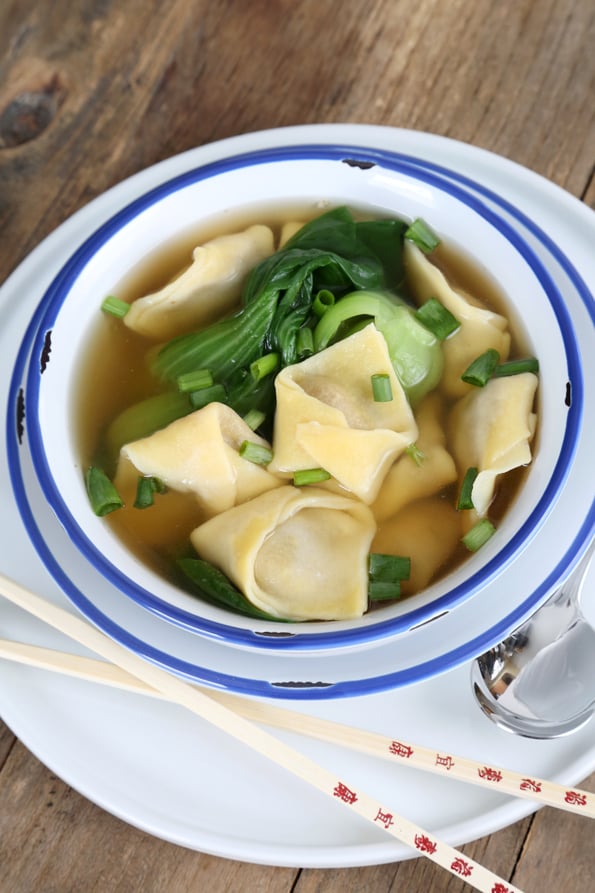 Light brown soup liquid with soup dumplings, chopped scallions, and baby bok choy in white bowl on white plate with chop sticks and a spoon