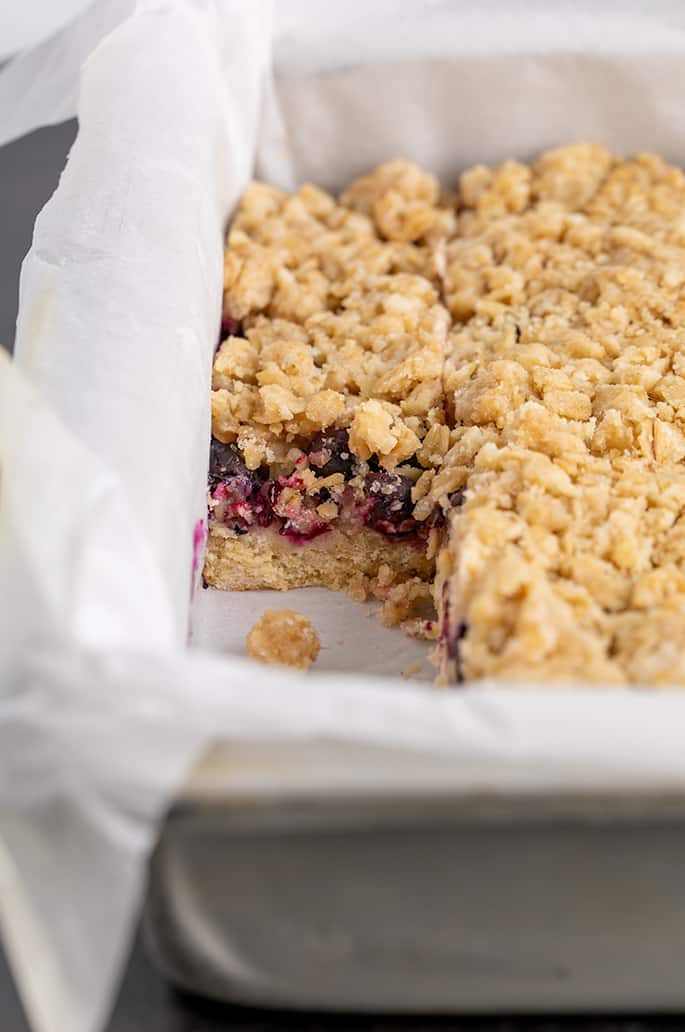 Pan of blueberry bars topped with crumble with one bar missing in metal pan with white liner