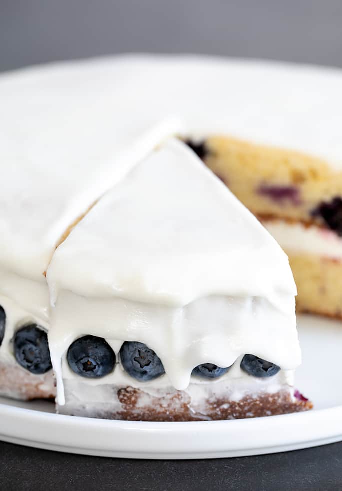 Gluten free lemon blueberry cake slice from behind, showing the glaze and fresh blueberries. 