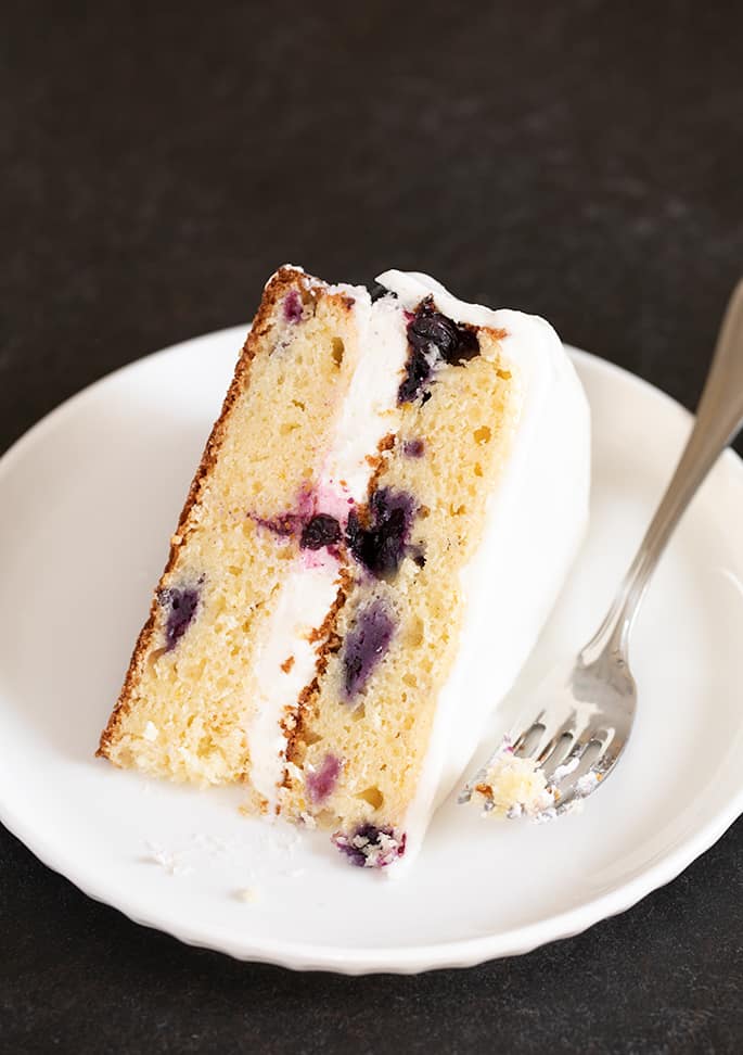 A piece of gluten free lemon blueberry cake with white frosting, on a plate.