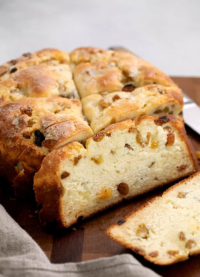 Partially sliced sweet flat round loaf of bread with raisins on brown cutting board with tan cloth