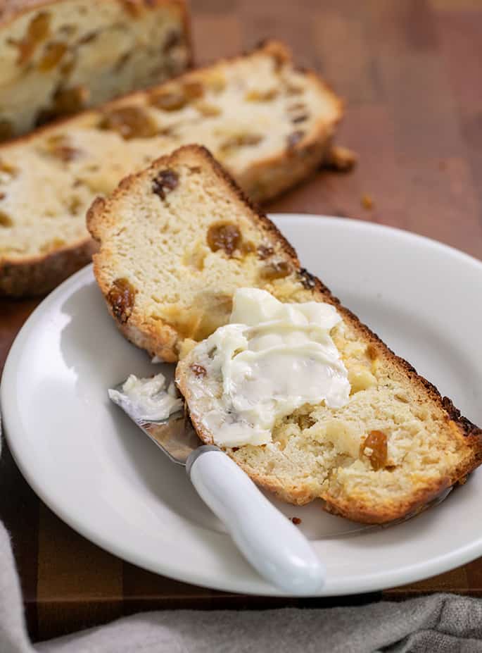 Slice of long narrow bread with yellow raisins on small white plate with butter and butter knife and more slices in background