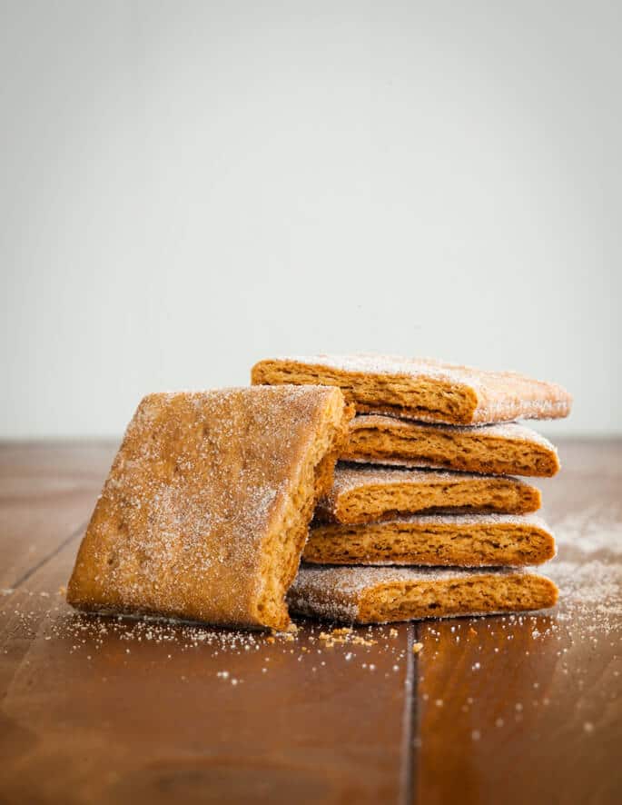 A stack of 5 halves of light brown graham crackers with one more half on the side of the stack on wooden brown slats with crumbs and sugar