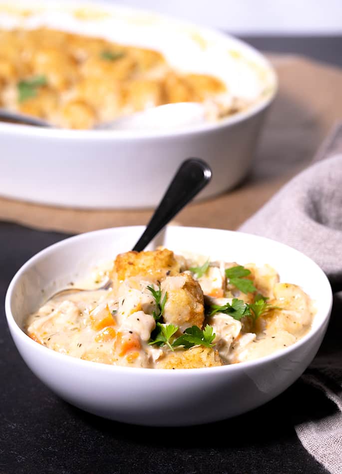 A bowl of chicken and dumplings with tater tots and a spoon