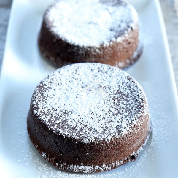 2 small round gluten free chocolate lava cakes with powdered sugar on top on rectangular white serving plate