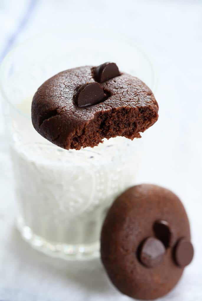 A chocolate flourless peanut butter cookie broken and sitting on the rim of a glass of milk