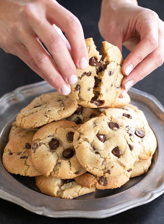 Hands breaking open a light brown cookie with chocolate chips held over a large round metal plate with many more cookies