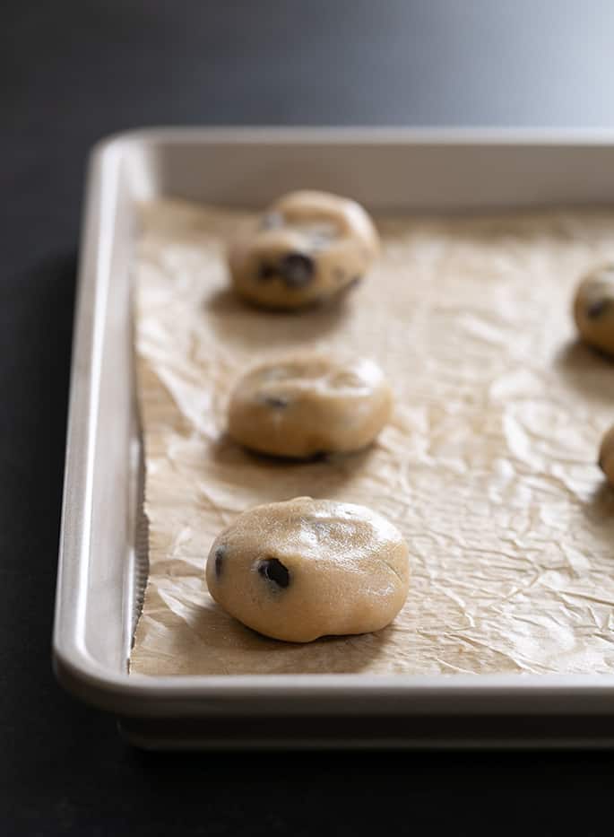 Raw disks of light brown dough with chocolate chips on a baking tray lined with brown paper