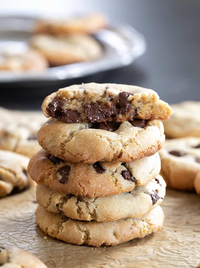 Stack of 5 gluten free chocolate chip cookies, with one broken open on top, on brown paper with more cookies in background