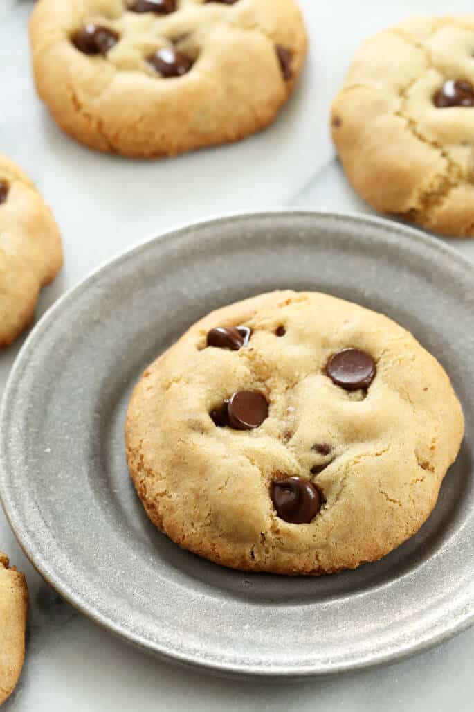 closeup image of a light brown cookie with chocolate chips on a small round metal plate on white cloth