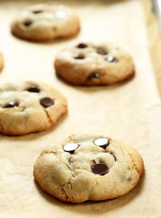 A batch of light brown cookies with chocolate chips baked on a tray lined in brown paper