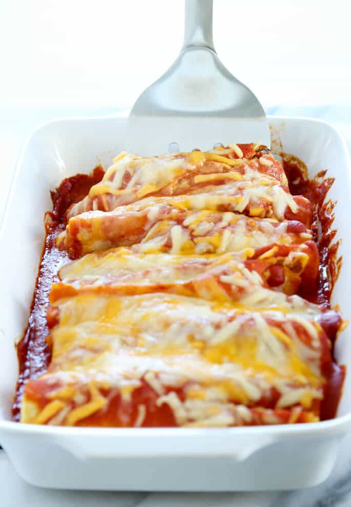 A white casserole dish with cheese on top of enchiladas and a spatula