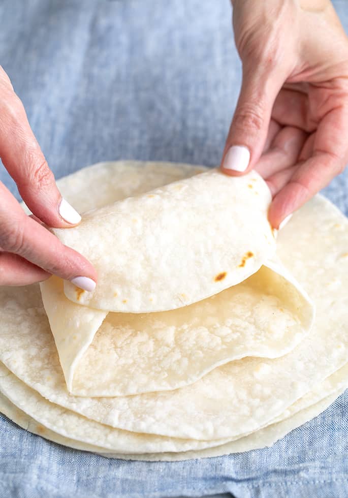 Hands folding top tortilla on a stack into a burrito shape, on blue cloth