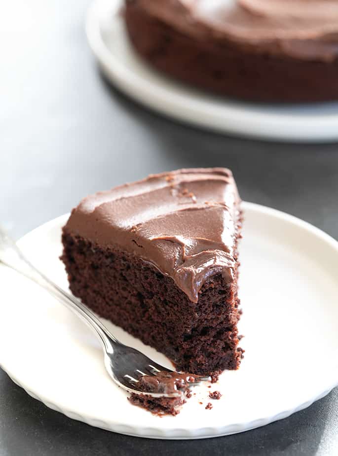 A piece of gluten free chocolate cake on a plate with a fork, with a soft brown frosting, and a bite taken.