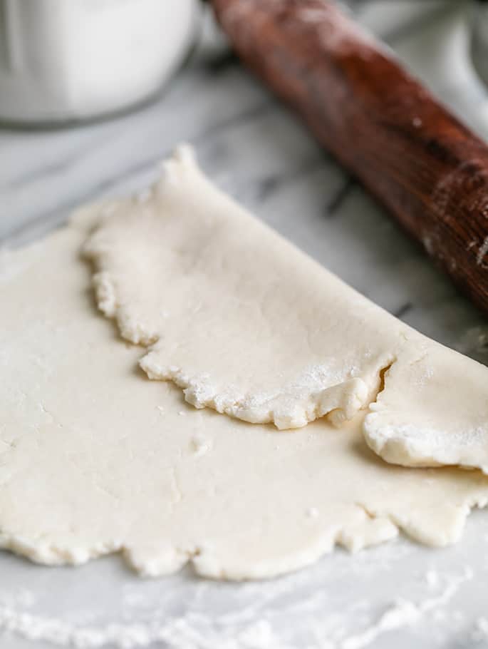 Raw flat tortilla dough folded over somewhat with a brown rolling pin and white jar in background on marble