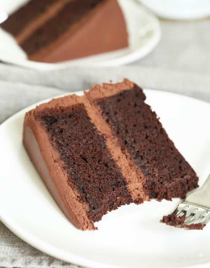 A piece of a chocolate cake with 2 layers and chocolate frosting in between on a white plate with a fork