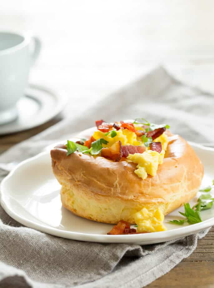 A white plate with a breakfast puff topped with eggs, bacon and herbs