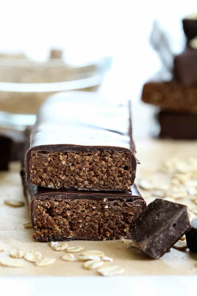 A stack of two brown rectangular homemade protein bars with white speckles and chocolate coating all around except for the bare ends, with scattered oats and chocolate chunks on brown paper