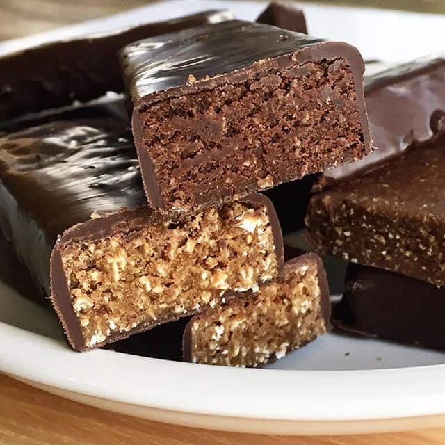 chocolate and peanut butter homemade protein bars covered in chocolate coating in a pile on white plate, with front 3 bars cut in half to reveal inside