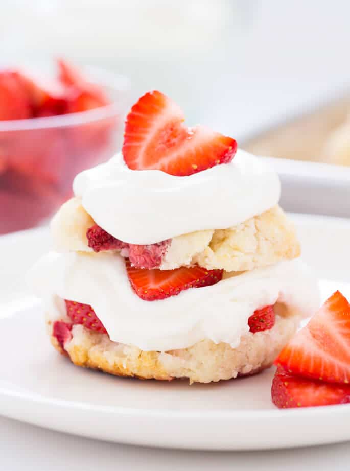 pale yellow and light brown strawberry shortcake biscuit sliced in half and layered with white whipped cream and more strawberries in the center and on top